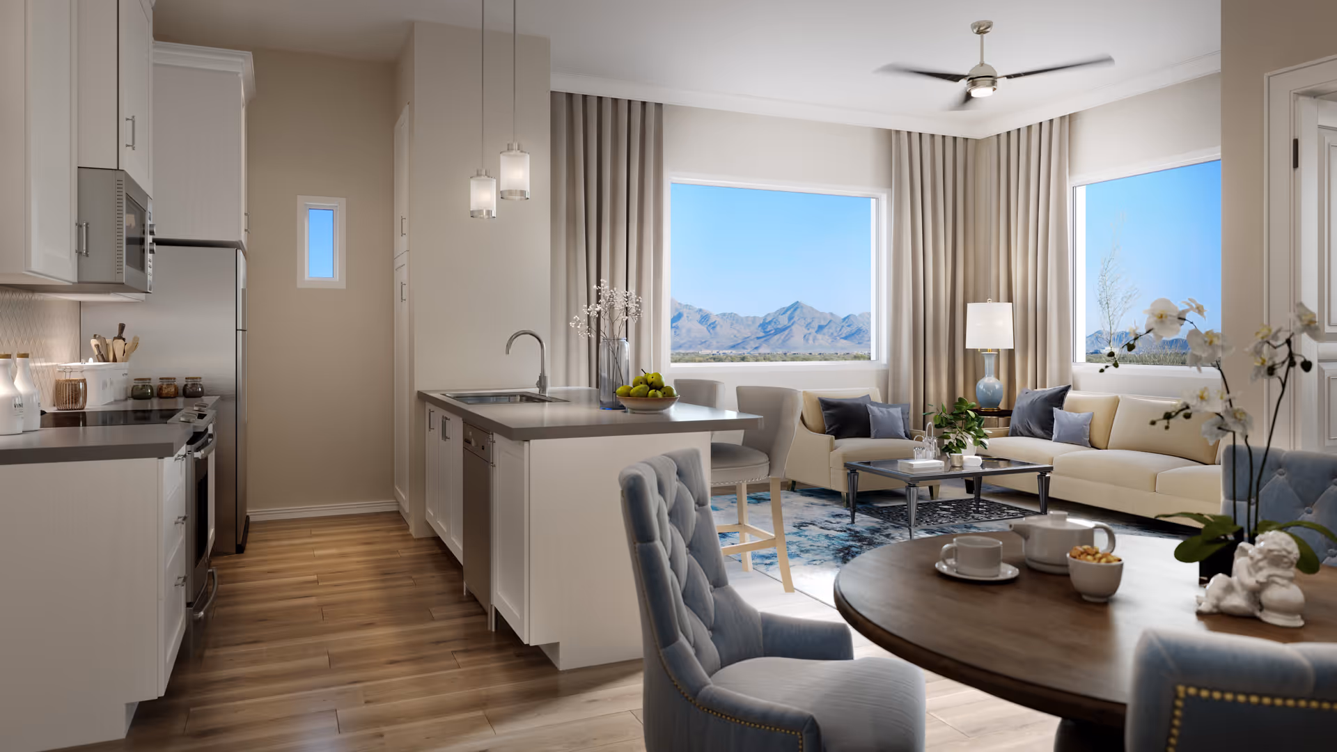 A bright and modern open-concept living space featuring a kitchen with white cabinets, stainless steel appliances, and a central island with a sink. Adjacent to the kitchen is a cozy living room with a beige sectional sofa, a coffee table, and large windows offering a scenic view of mountains. In the foreground, there is a round wooden dining table with blue upholstered chairs and a white orchid centerpiece.