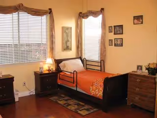 A cozy bedroom in a senior living facility with a bed, dresser, and windows.