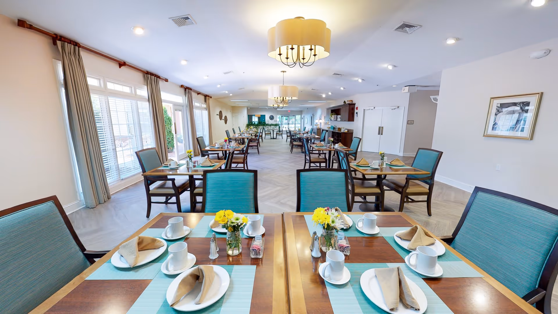 A bright and spacious dining room with multiple wooden tables set with white cups, plates, beige napkins, and small flower vases. The room has large windows with beige curtains allowing natural light to fill the space. Blue cushioned chairs surround the tables, and modern ceiling lights hang overhead.
