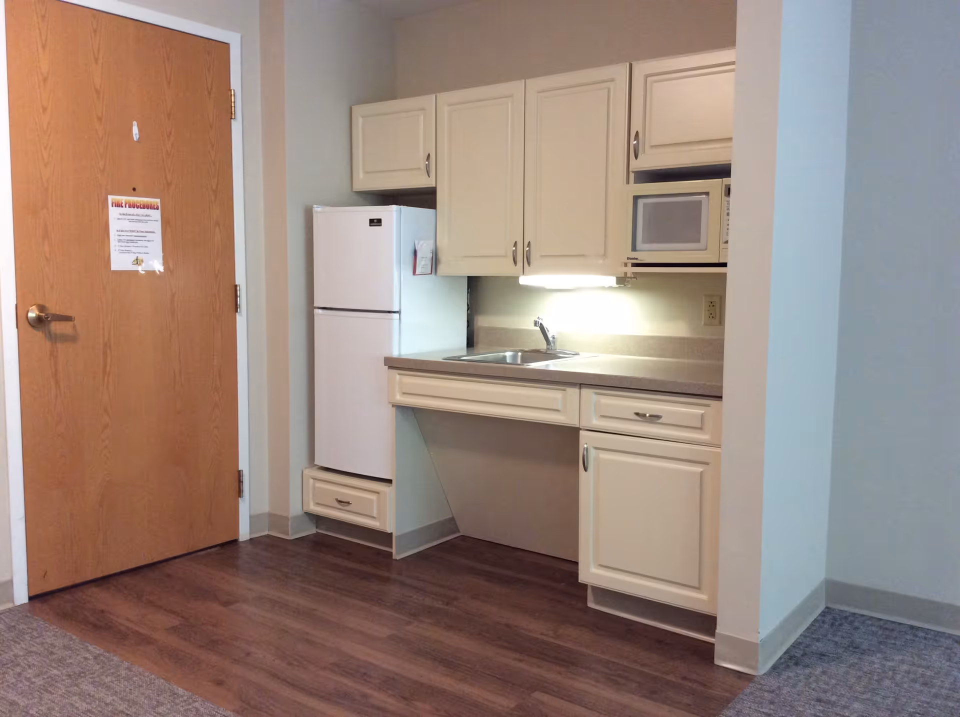 Small kitchenette area with white cabinets, a white refrigerator, a microwave, a sink, and a wooden door on the left side. The floor is a combination of wood and carpet.