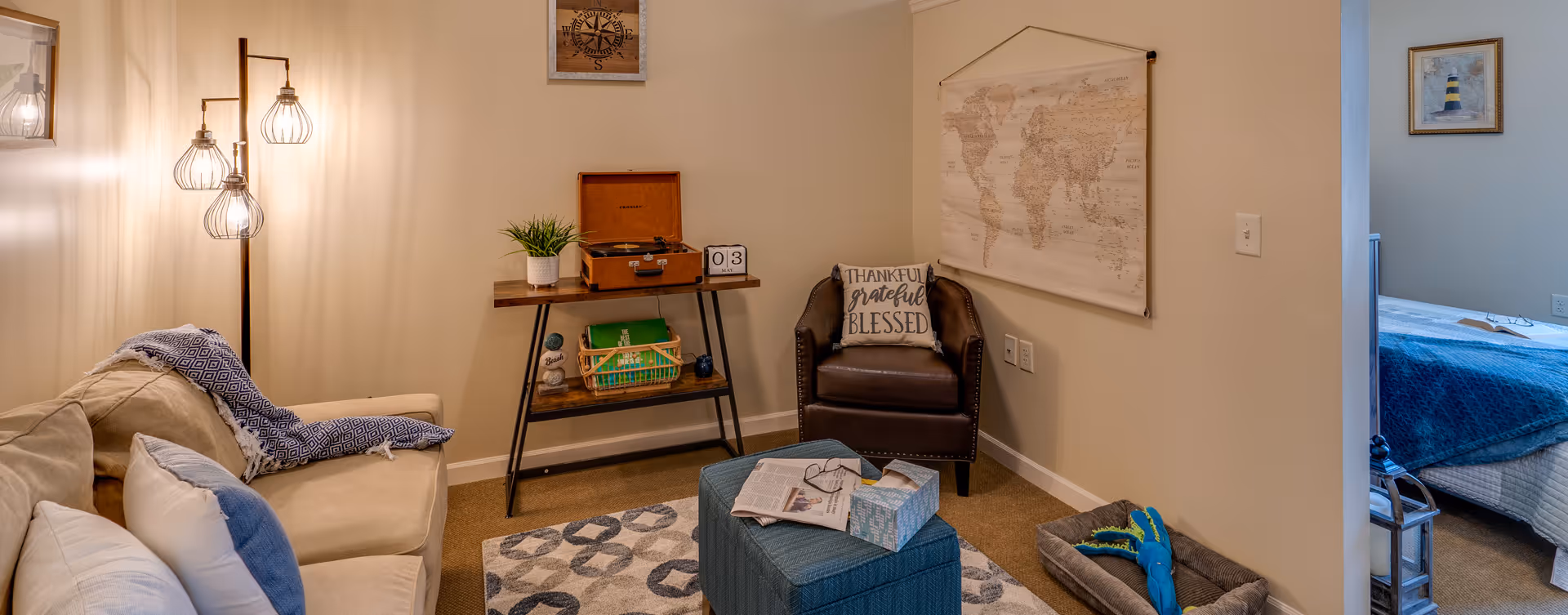 Cozy living room area with a beige couch adorned with pillows and a blue patterned throw blanket. A modern floor lamp with three exposed bulbs stands beside the couch. A wooden console table holds a potted plant, a vintage-style record player, and a calendar block showing May 3. A brown leather armchair with a pillow that reads 'Thankful grateful blessed' is positioned near a wall hanging of a world map. A blue ottoman with a newspaper, glasses, and a tissue box sits on a patterned rug. A pet bed with a blue toy is on the floor. In the background, a bedroom with a blue bedspread and framed artwork is partially visible through an open doorway.
