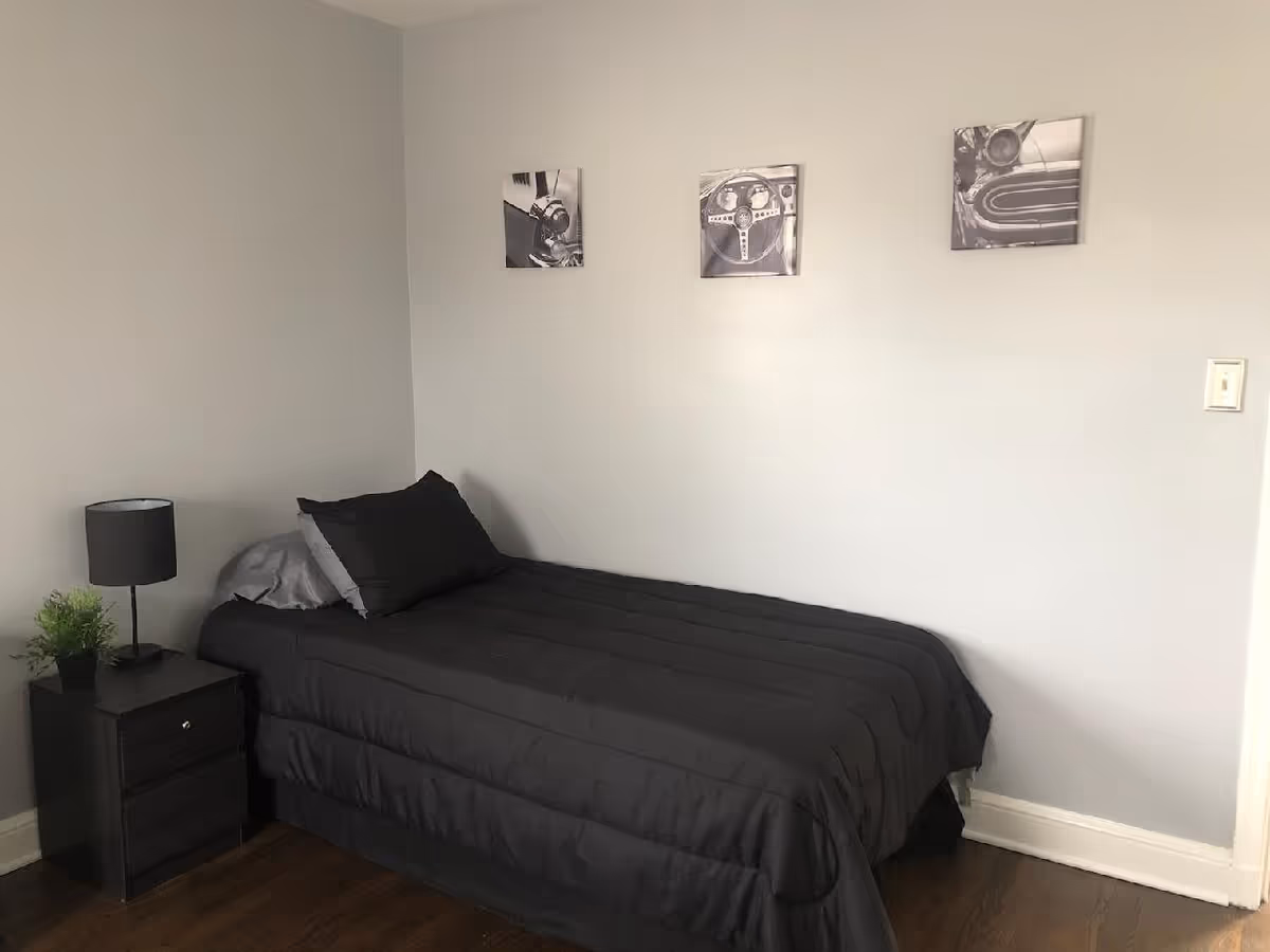 A small bedroom with a single bed covered in black bedding and pillows. Next to the bed is a black nightstand with a small green plant and a black lamp. Three black and white framed pictures are hung on the light gray wall above the bed. The floor is wooden.