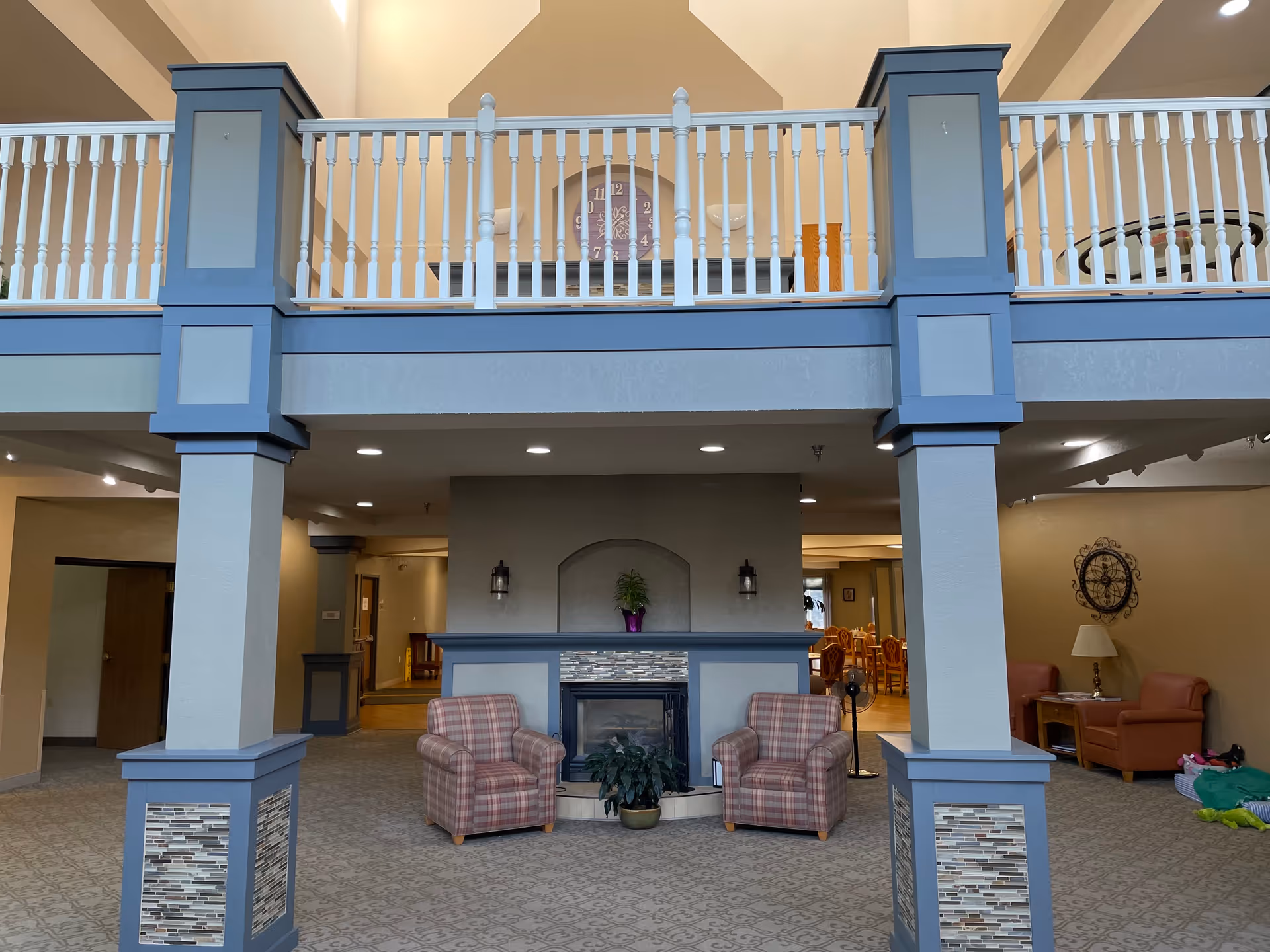 Interior view of a senior living facility common area with two plaid armchairs facing a fireplace. The room features blue and gray pillars with decorative tile accents, a balcony railing above, and a clock on the wall behind the fireplace. Additional seating and tables are visible in the background.