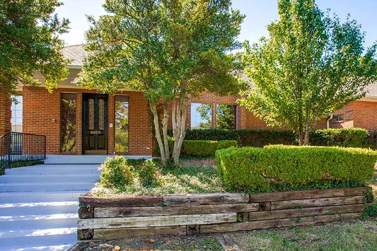 Front exterior view of a brick building with a black door numbered 6601, surrounded by green trees and neatly trimmed bushes, with concrete steps leading up to the entrance.