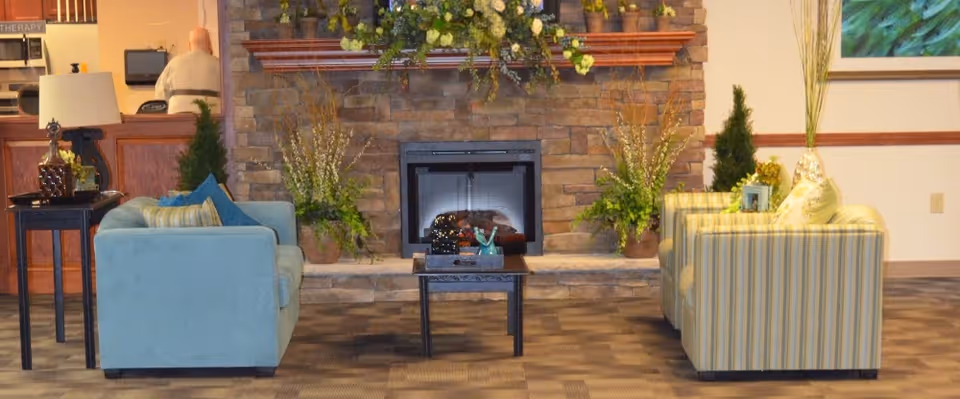 A cozy living room area with a stone fireplace in the center, flanked by two sofas—one light blue and one striped. There are decorative plants on either side of the fireplace and a small coffee table in front of it. A lamp and other decor items are on a side table next to the blue sofa. In the background, a person is seated at a counter or desk area.