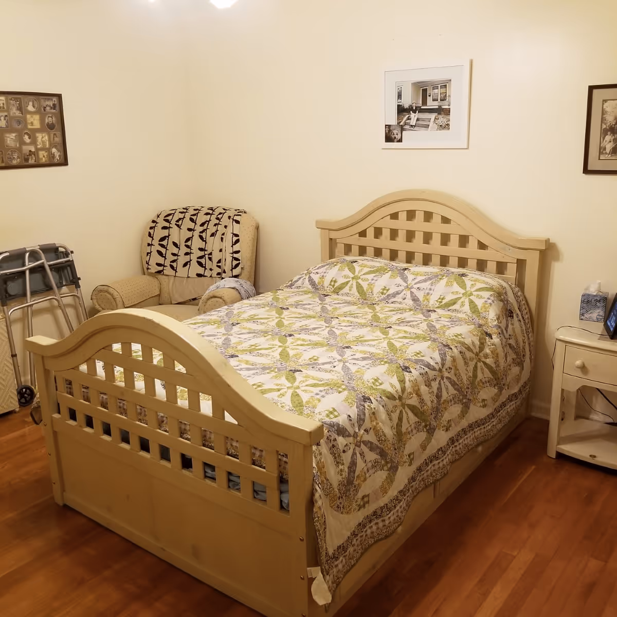 A cozy bedroom with a wooden bed frame and a quilted bedspread featuring a green and purple pattern. There is a beige armchair with a patterned blanket draped over it in the corner, a walker next to the chair, and a small white nightstand with a tissue box and a framed photo. The walls are decorated with framed pictures.