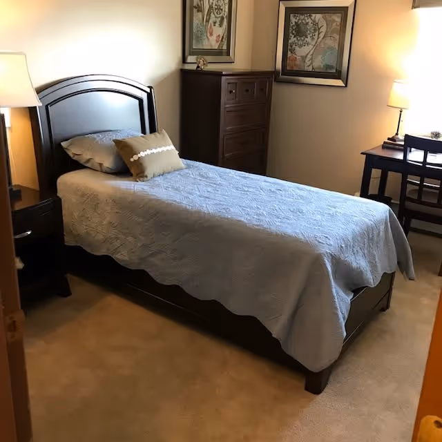 A neatly made single bed with a light blue quilt and two pillows, one gray and one brown with a white pattern, in a cozy bedroom. The room features a wooden nightstand with a lamp on the left side of the bed, a tall wooden dresser against the wall, two framed floral artworks hanging above the dresser, and a small desk with a chair and lamp near the window on the right.