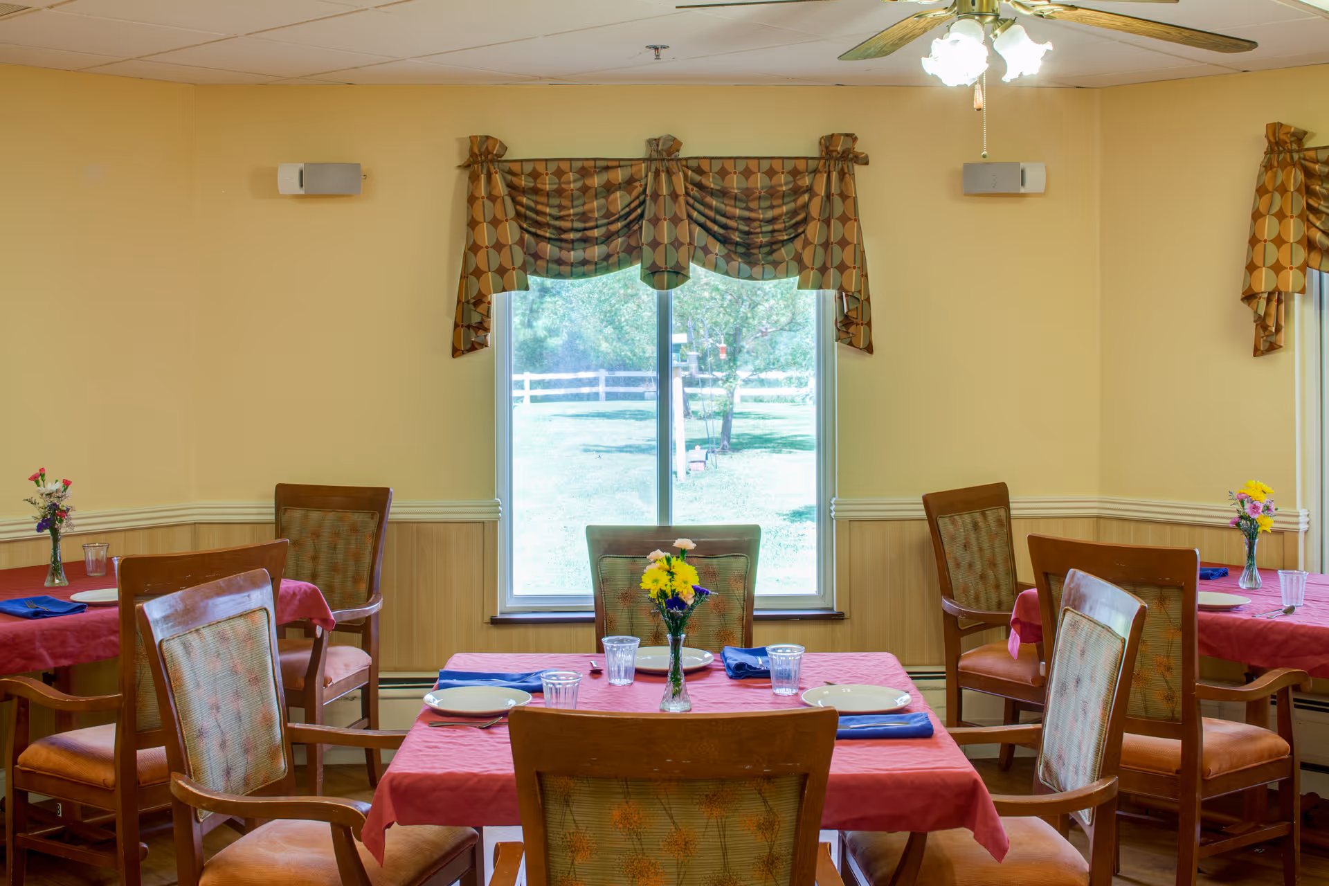 A dining room with several wooden tables covered in red tablecloths, each set with plates, glasses, and blue napkins. There are wooden chairs with cushioned seats around the tables. A window with patterned valances shows a green outdoor area with trees and a white fence. The room has beige walls with wood paneling and a ceiling fan with lights.