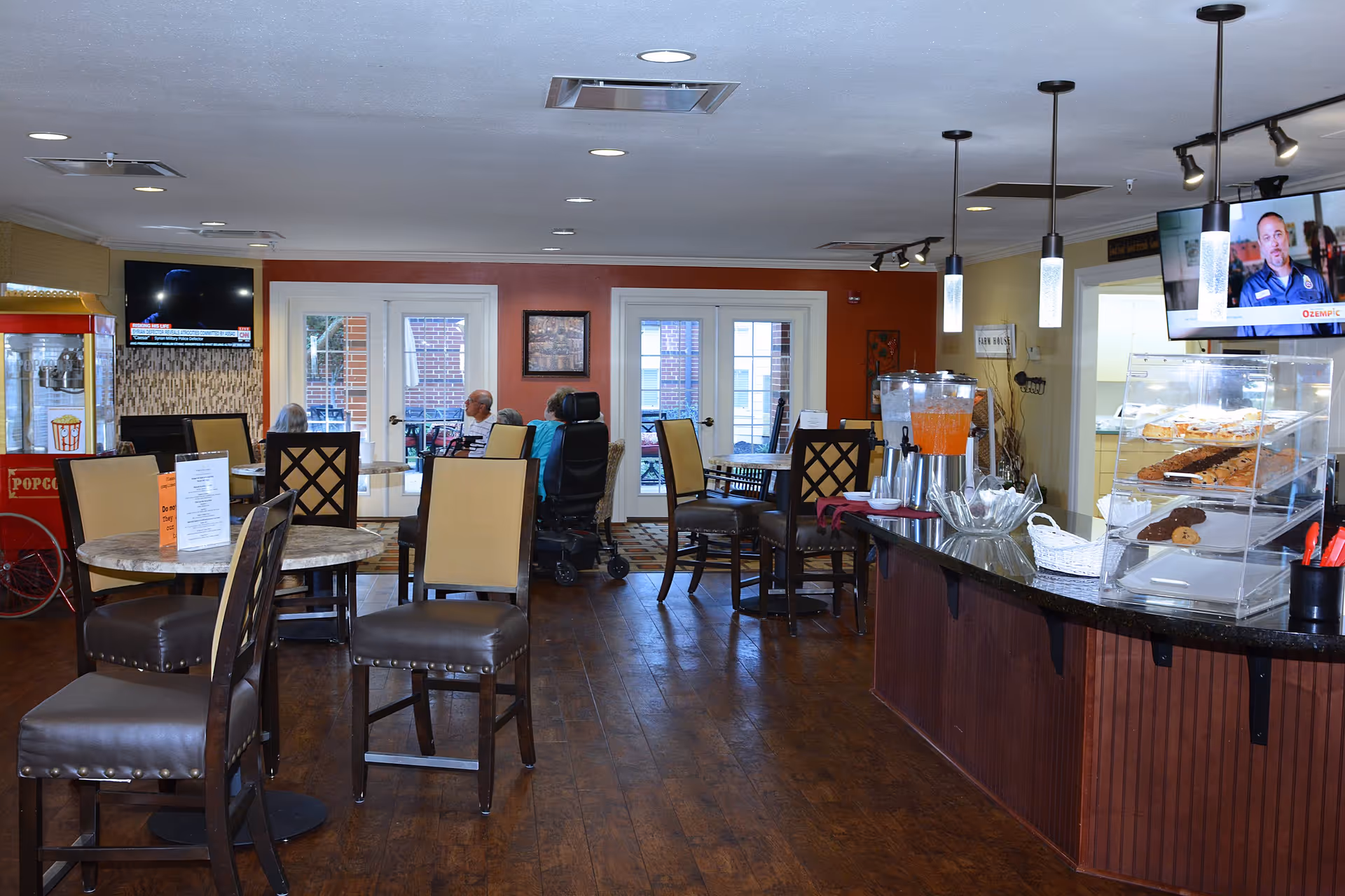 Interior view of a dining area in a senior living facility with several tables and chairs, a counter displaying baked goods and a beverage dispenser, a popcorn machine, and a television mounted on the wall. There are a few elderly people seated at the tables near the windows.