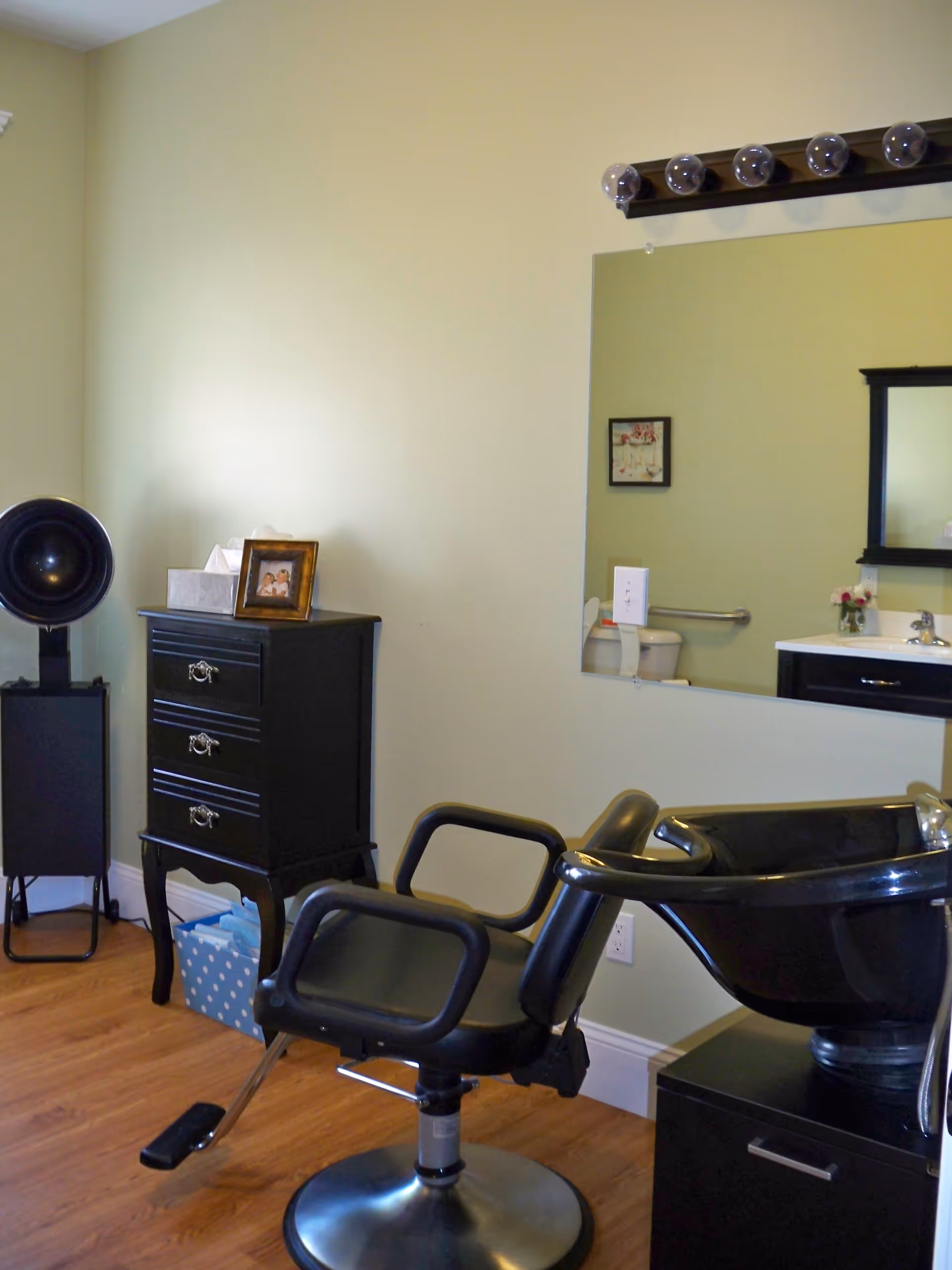 Interior view of a salon area in a senior living facility with a black salon chair and a black hair washing station. There is a black chest of drawers with a framed photo and tissue box on top, a large mirror on the wall reflecting a sink and toilet with grab bars, and a wooden floor.