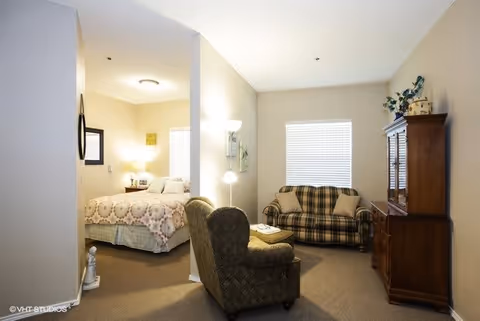 A cozy living space in a senior living facility featuring a small sitting area with a patterned armchair, a plaid loveseat, a wooden cabinet with decorative items on top, and a separate bedroom area with a bed covered in a quilted bedspread. The room is softly lit with natural light coming through a window with blinds and a floor lamp near the armchair.
