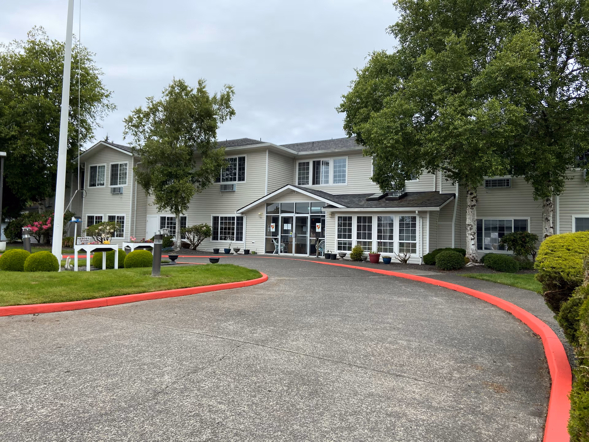 Two-story beige senior living building with a covered main entrance, curved driveway, and landscaped lawn with trees.