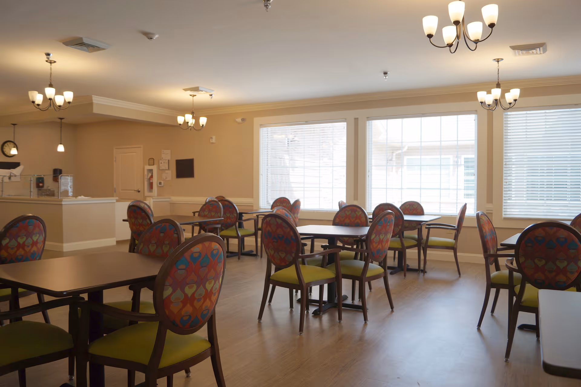 Communal dining room with round tables and colorful patterned chairs under chandeliers and large windows.