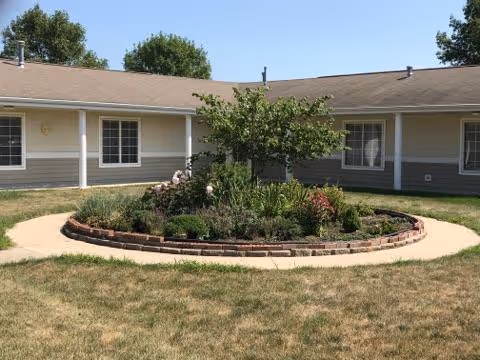 A circular garden bed with various plants and flowers in the center of a paved walkway, surrounded by a single-story building with beige siding and white trim under a clear blue sky.