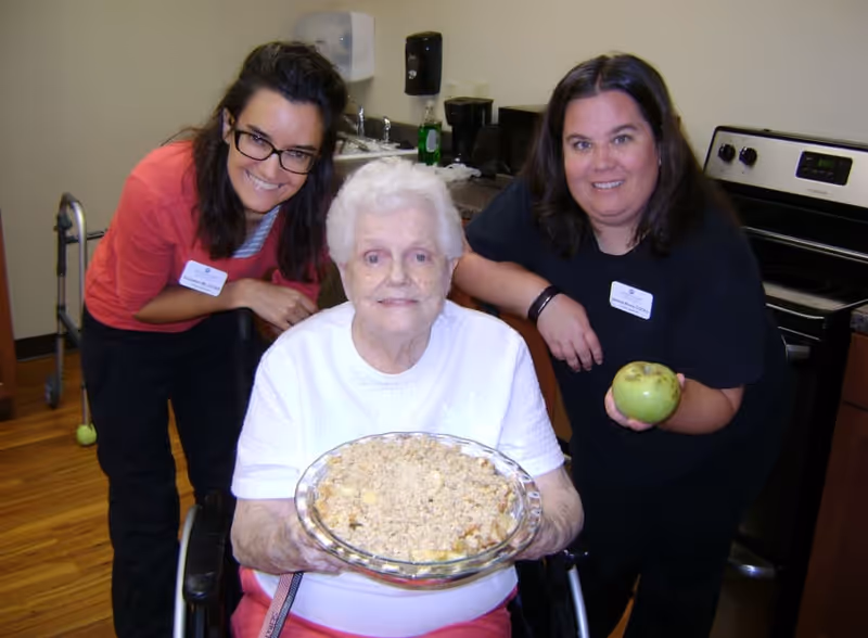 An elderly woman in a wheelchair holding a glass dish with a baked dessert, flanked by two women standing behind her in a kitchen setting. One woman is holding a green apple, and both women are smiling. The kitchen includes a stove, sink, and countertop with various items.