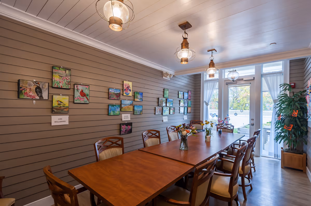 Communal dining room with a long wooden table and chairs, floral centerpieces, artwork on a paneled wall, and glass doors to the outside.