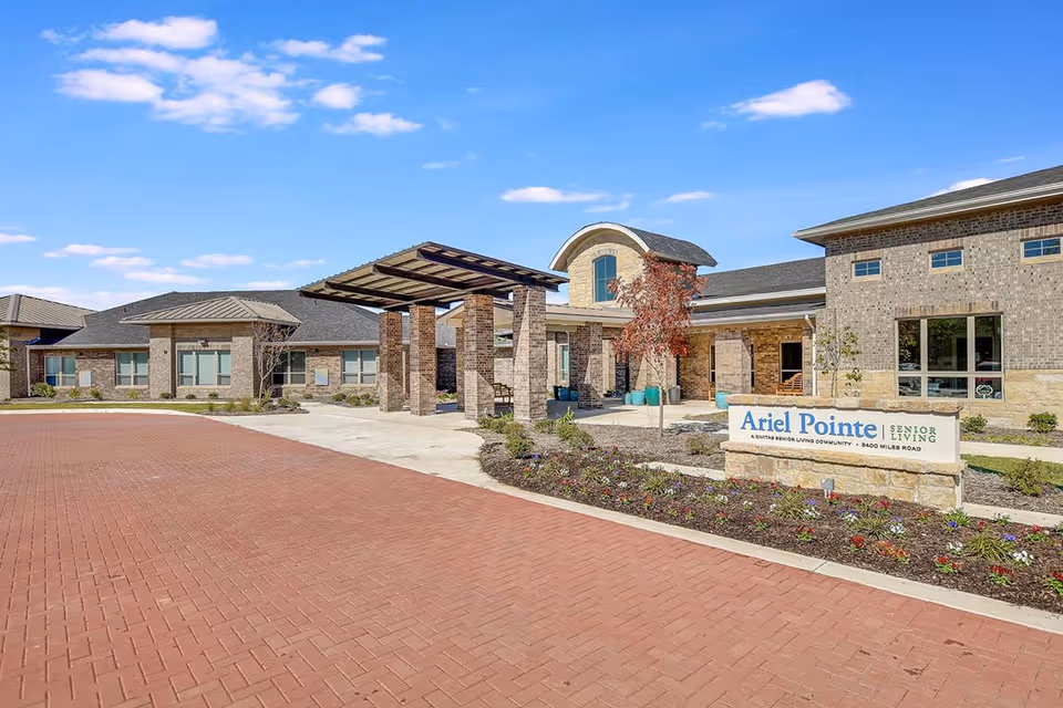 Front exterior view of Ariel Pointe Of Sachse senior living facility with a brick driveway, landscaped flower beds, and a covered entrance supported by brick pillars under a blue sky with scattered clouds.