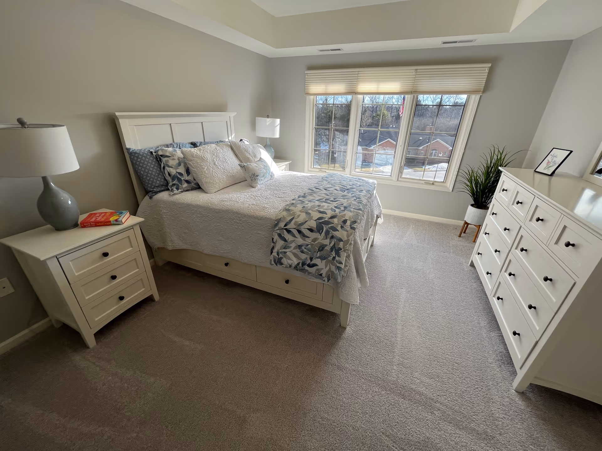 A bright and neatly arranged bedroom with a white bed frame and bedding featuring blue and white patterned pillows and a matching throw. There is a white nightstand with a lamp and books on the left side of the bed, and a large white dresser with multiple drawers on the right side of the room. A large window with blinds lets in natural light, and a potted plant is placed near the dresser. The room has light gray walls and beige carpet flooring.