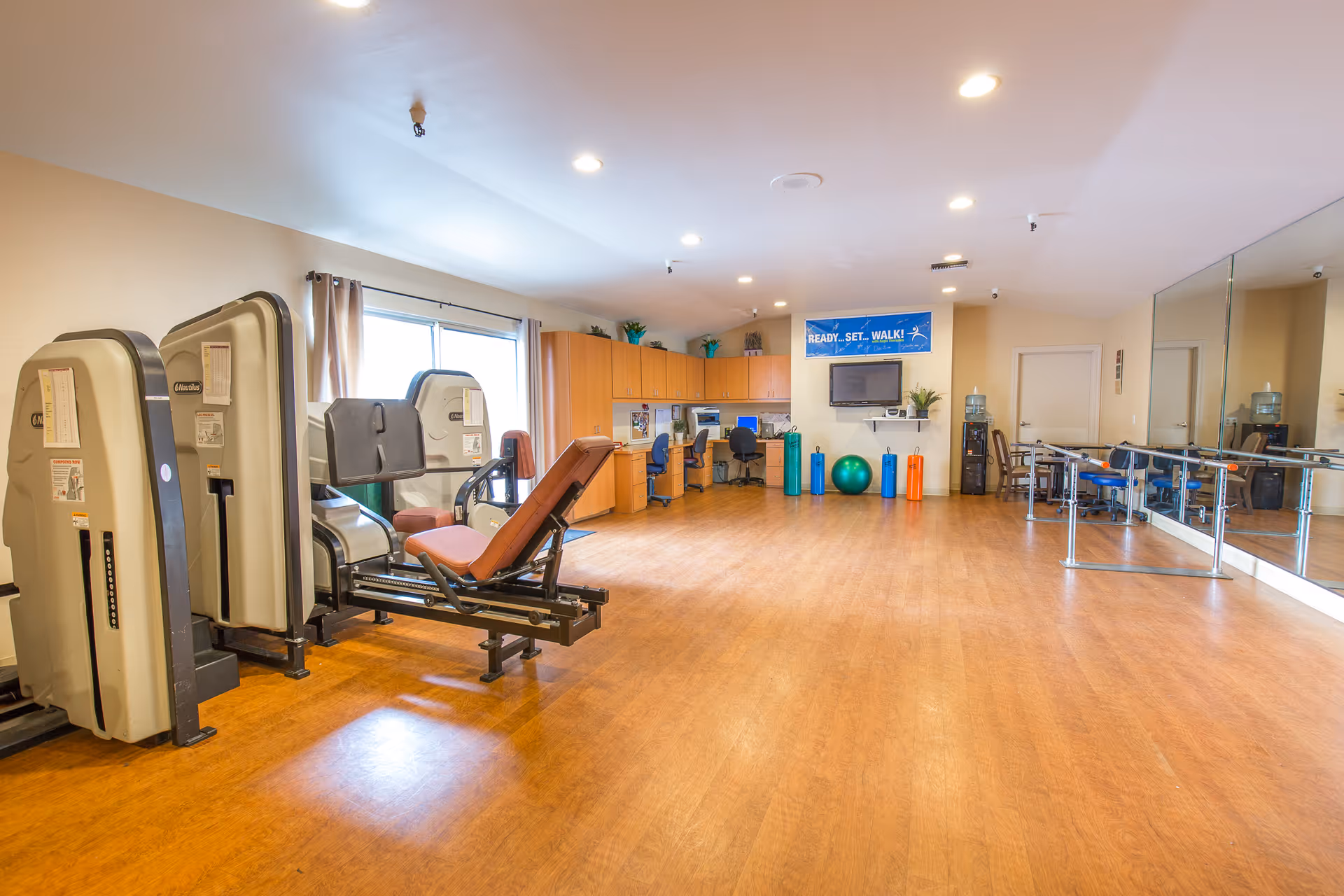 A spacious exercise room with wooden flooring, exercise machines, a large mirror on one wall, a desk with chairs, and various exercise equipment including stability balls and foam rollers. A banner on the wall reads 'READY, SET, WALK!'.