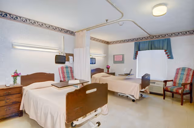A shared patient bedroom with two hospital-style beds, recliner chairs, bedside tables, a privacy curtain, and a window.