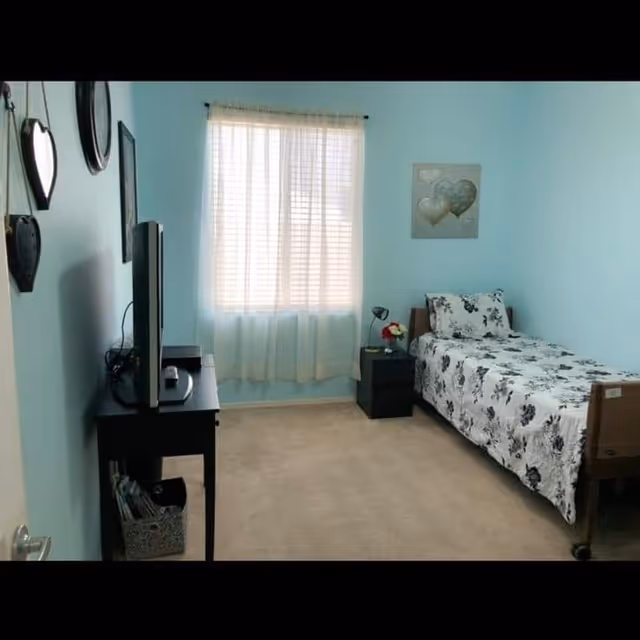 A small bedroom with light blue walls, a single bed with floral bedding, a black nightstand with a lamp and flowers, a window with sheer white curtains, a black TV stand with a flat-screen TV, and decorative wall hangings.