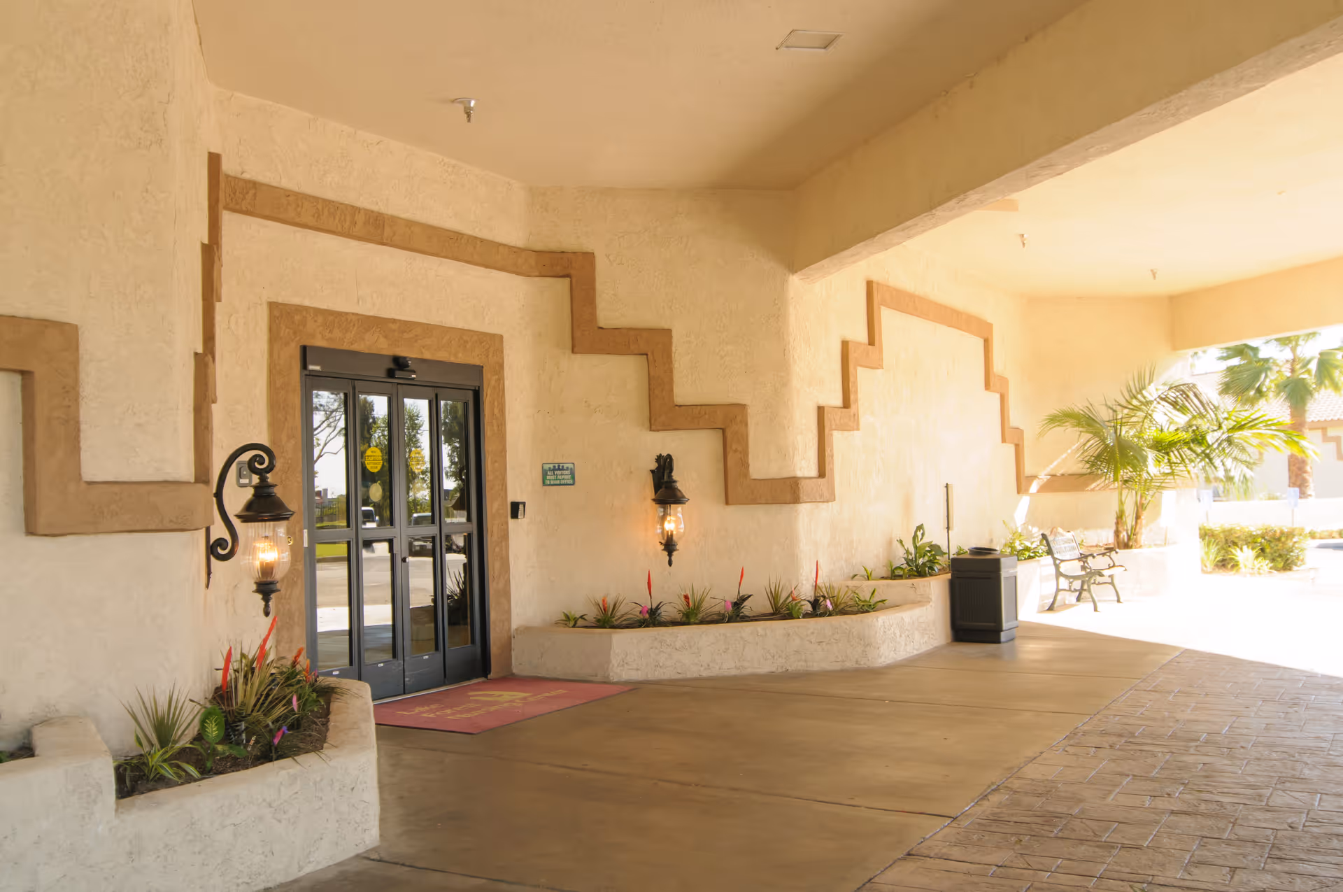 Covered building entrance with glass double doors, decorative stucco walls, planter beds, wall lanterns and a bench.