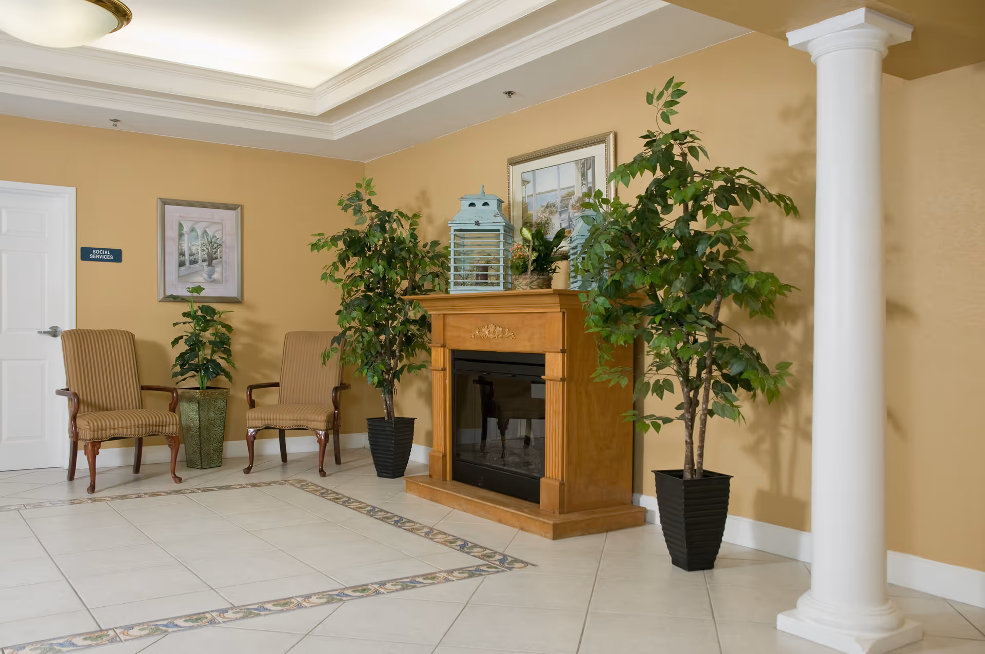 Reception/lobby area with two striped armchairs, a wooden fireplace mantel, potted plants, and a white decorative column.