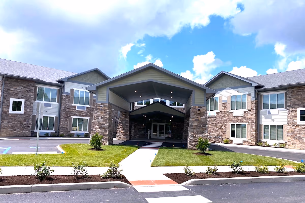 Front exterior view of a two-story assisted living facility with stone and siding facade, a covered entrance with pillars, landscaped grass and bushes, and a partly cloudy blue sky.