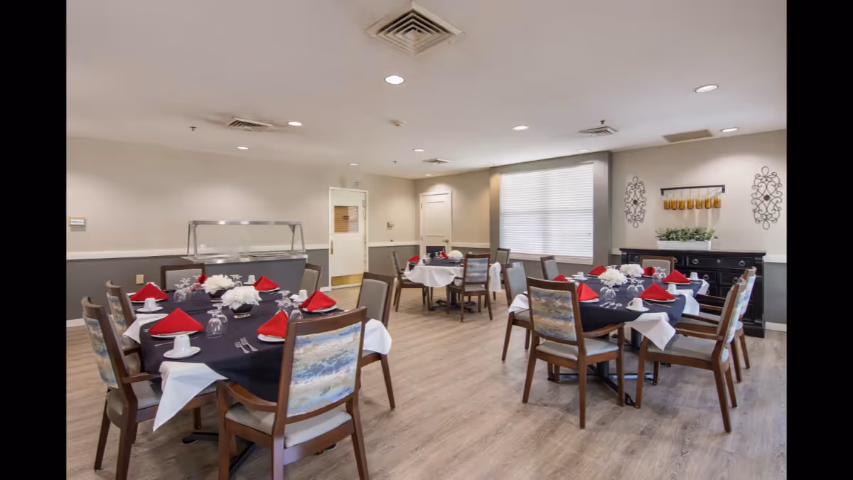 A dining room in a senior living facility with multiple round tables set with black and white tablecloths, red folded napkins, glassware, and white floral centerpieces. The room has wooden chairs with patterned backs, light wood flooring, neutral-colored walls, a window with blinds, and decorative wall art. A serving station is visible against one wall.