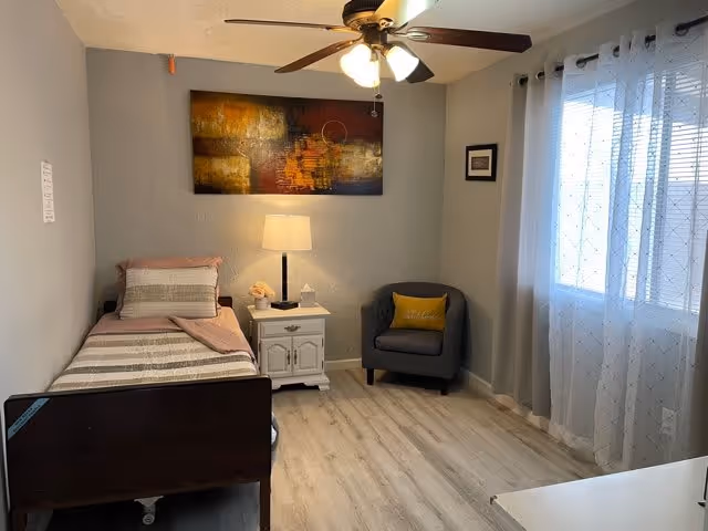 A small, neatly arranged bedroom with a single bed on the left side, covered with striped bedding and a pink blanket. Next to the bed is a white nightstand with a lamp, a small decorative item, and a tissue box. Above the nightstand hangs an abstract painting with warm colors. A gray armchair with a yellow pillow is positioned near a window with sheer white curtains. The room has light wood flooring, light gray walls, and a ceiling fan with lights.