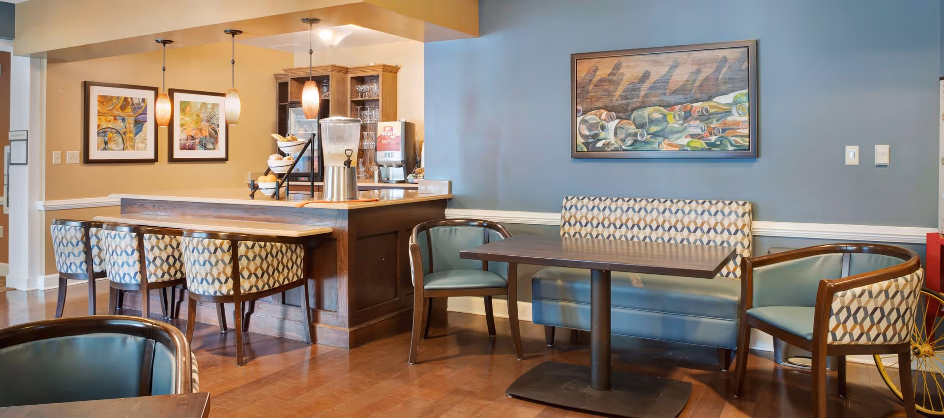 A cozy dining area with a wooden table, patterned cushioned chairs, and a bench against a blue wall. The space includes a counter with four high chairs, pendant lights hanging above, and framed artwork on the walls. There is a beverage dispenser and a coffee machine on the counter.