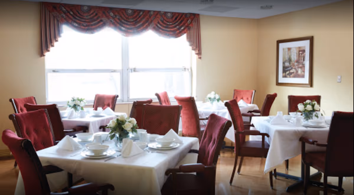 Dining room with several tables set with white tablecloths, red upholstered chairs, and floral centerpieces beneath a large window.