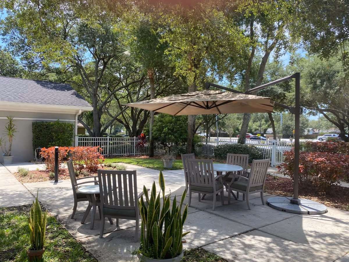 Outdoor patio area with a round table and four chairs under a large beige umbrella. The patio is surrounded by greenery including trees, bushes, and potted plants. A white fence and a building with a gray roof are visible in the background under a clear blue sky.