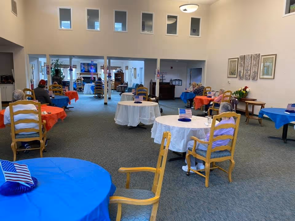 A spacious dining area in a senior living facility with round tables covered in red, white, and blue tablecloths. Each table has a small American flag centerpiece. Several elderly residents are seated at the tables, spaced apart. The room has high ceilings, light-colored walls, and several small windows near the ceiling. In the background, there is a television and additional seating areas.