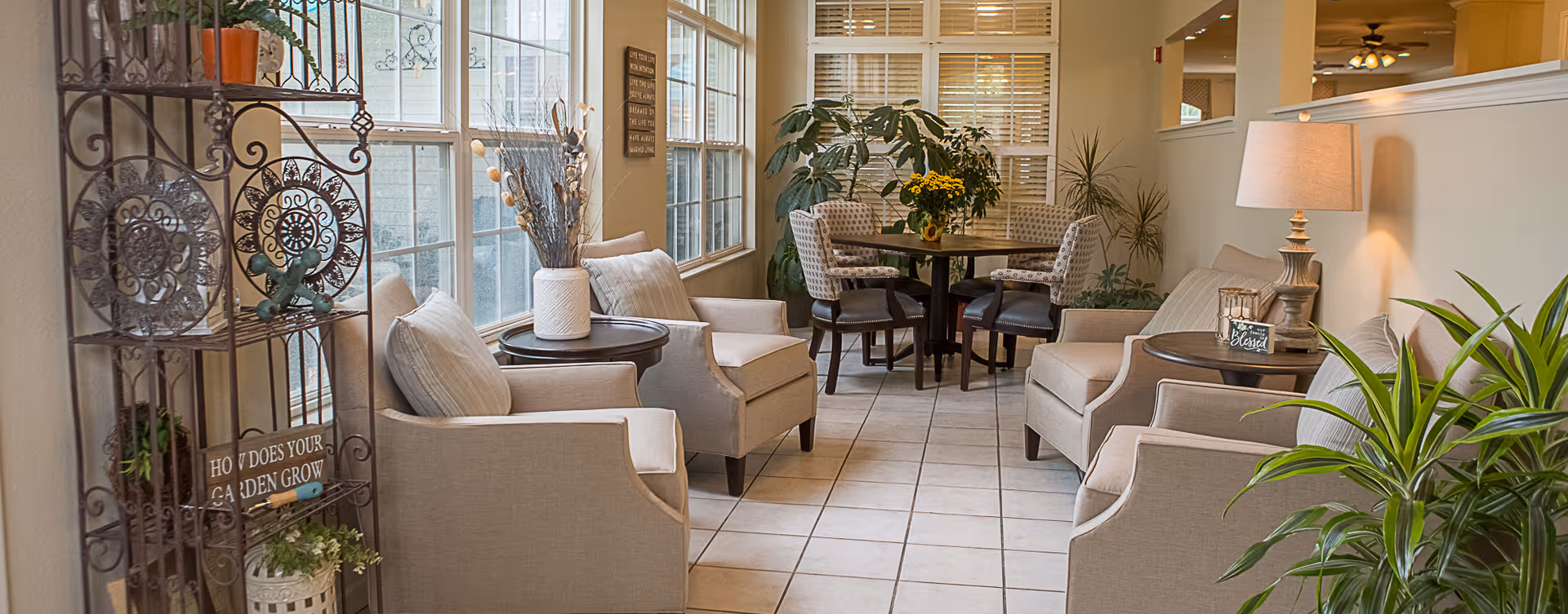 Bright communal seating area with several upholstered armchairs, side tables, plants and a small round dining table by large windows.