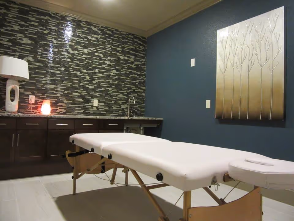A massage or therapy room with a white massage table in the center. The room has dark wooden cabinets with a granite countertop, a modern mosaic tile backsplash, a white lamp, and a small glowing salt lamp on the counter. The walls are painted dark blue, and there is a decorative metallic wall art piece featuring tree designs.