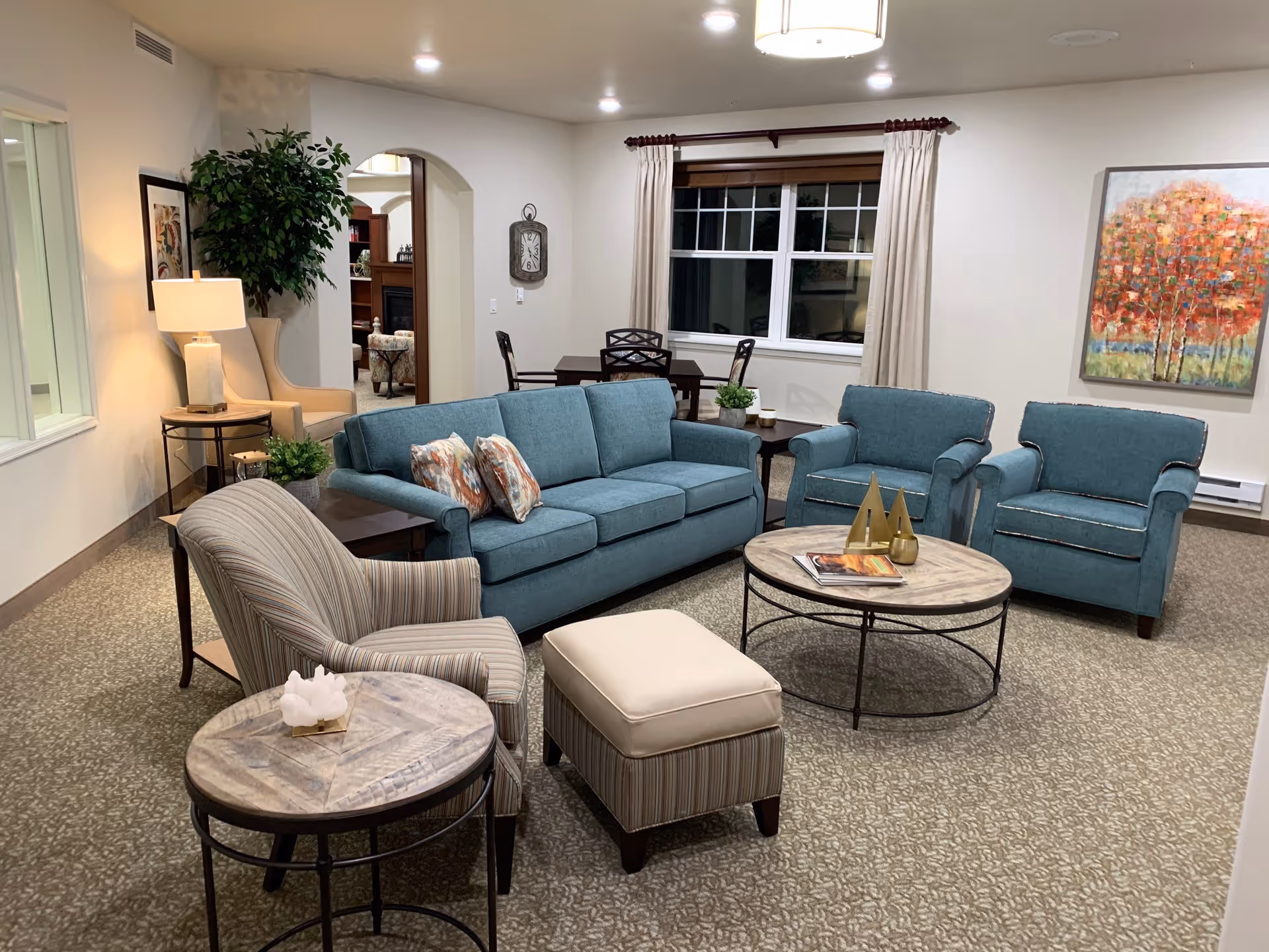 A cozy living room with a blue sofa, two matching blue armchairs, a striped armchair, and a beige ottoman. There are two round wooden coffee tables, one with decorative items and magazines. The room has a large window with beige curtains, a colorful painting on the wall, a floor lamp, and a potted plant. In the background, there is a dining table with chairs and an arched doorway leading to another room.