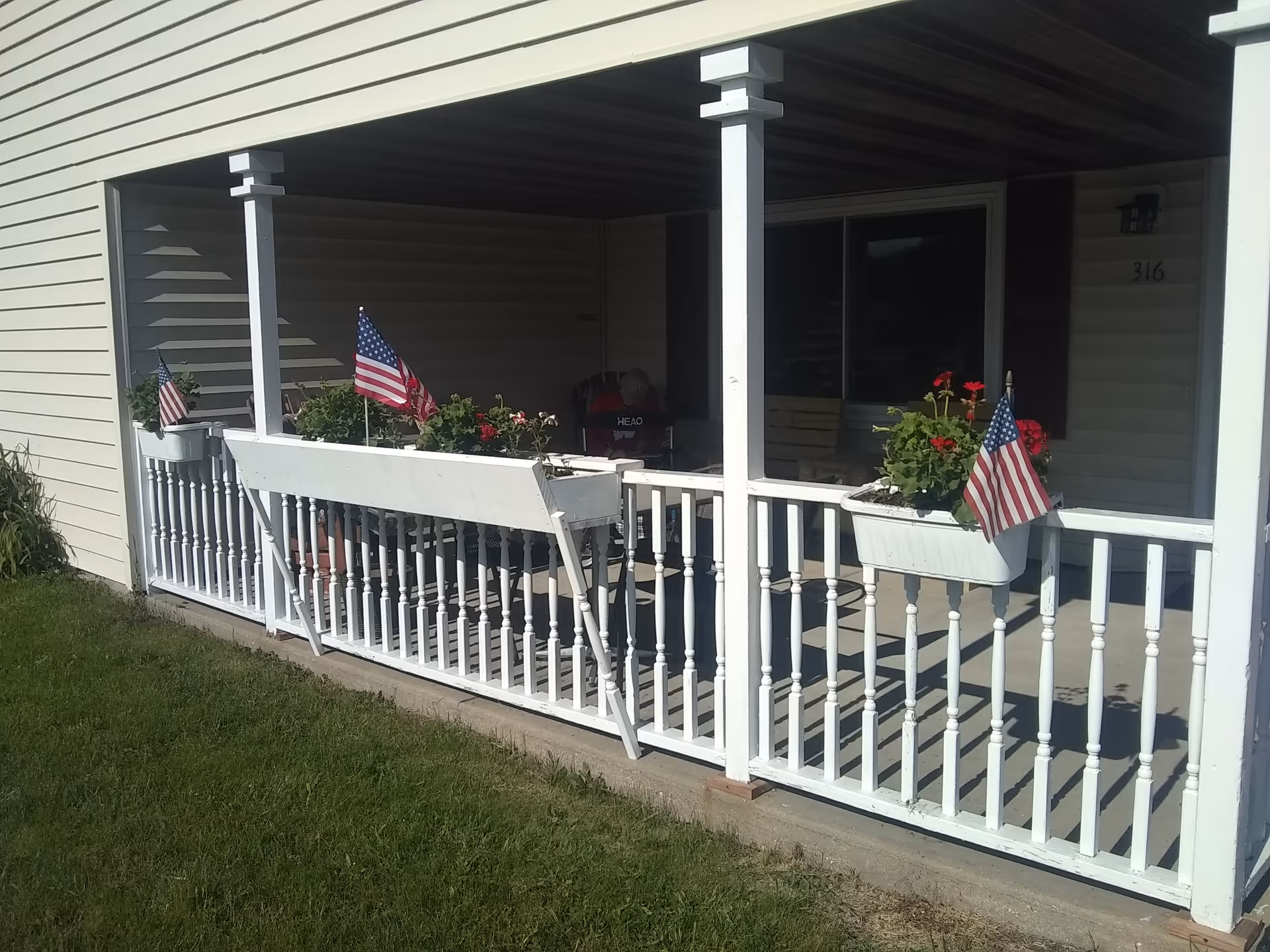 Covered porch area with white railing and flower boxes containing red flowers and American flags. There is a chair and a bench inside the porch, and the porch is attached to a beige building with the number 316 visible near the door.