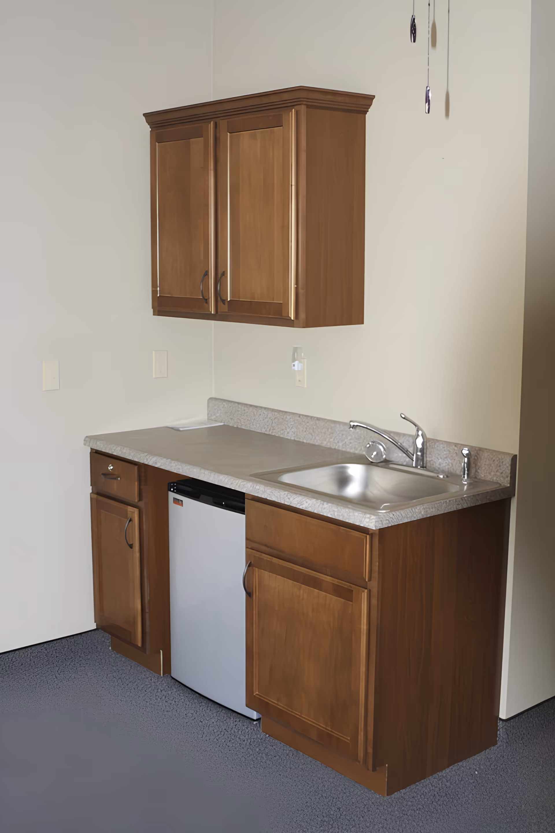 A small kitchenette area with wooden cabinets, a countertop with a stainless steel sink and faucet, and a compact refrigerator underneath the counter. The walls are light-colored and the floor has a textured surface.
