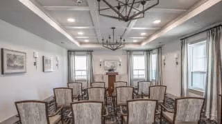 A well-lit room with multiple upholstered chairs arranged in rows facing a fireplace with a mirror above it. The room features large windows with curtains, a coffered ceiling with chandeliers, and framed artwork on the walls.