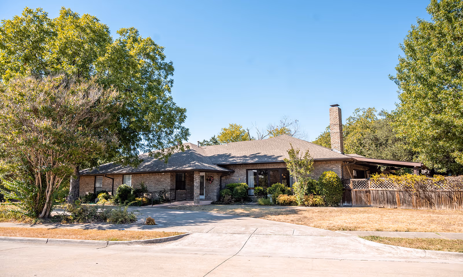 Exterior view of a single-story brick building surrounded by trees and shrubs under a clear blue sky. The building has a sloped roof, a chimney, and a wooden fence on the right side. There is a paved driveway and a sidewalk in front of the building.