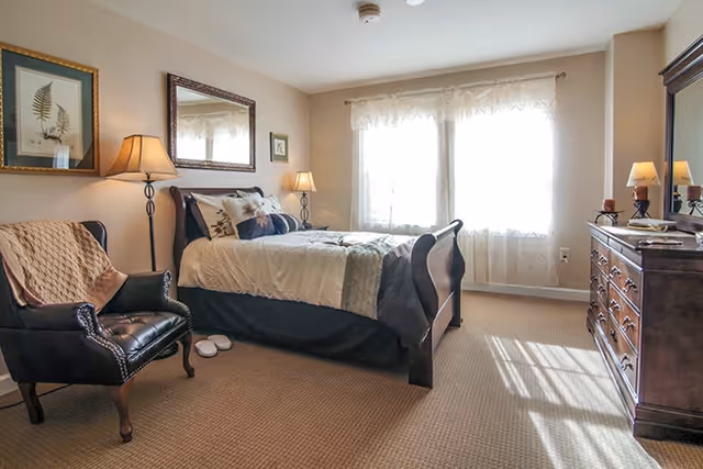 A well-lit bedroom with a large bed covered in white and beige bedding, a dark wooden headboard and footboard, a black leather armchair with a beige throw, two lamps on either side of the bed, a dresser with a mirror, and two windows with sheer white curtains letting in natural light.