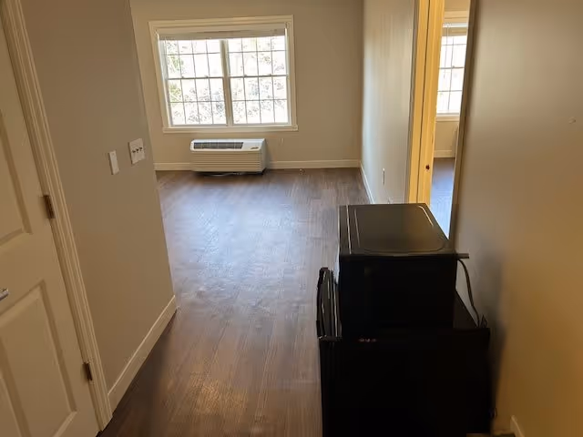 View down a hallway into a room with large windows and a wall-mounted air conditioning unit below the windows. The floor is wood-style laminate, and there is a microwave and mini refrigerator stacked on the right side of the hallway. The walls are painted beige.