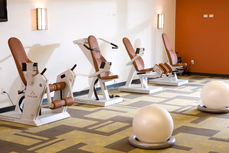A fitness room with four exercise machines lined up against a white wall with brown padded seats and handles. Two large exercise balls are placed on the patterned floor in the foreground. The room has a warm color scheme with an orange accent wall and wall-mounted lights.