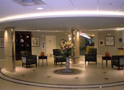 A spacious, well-lit common area with a circular design featuring a central round table with a large floral arrangement. Surrounding the table are several upholstered chairs and small side tables. The room has beige walls, decorative columns, framed artwork, and a built-in wooden cabinet with shelves displaying decorative items.