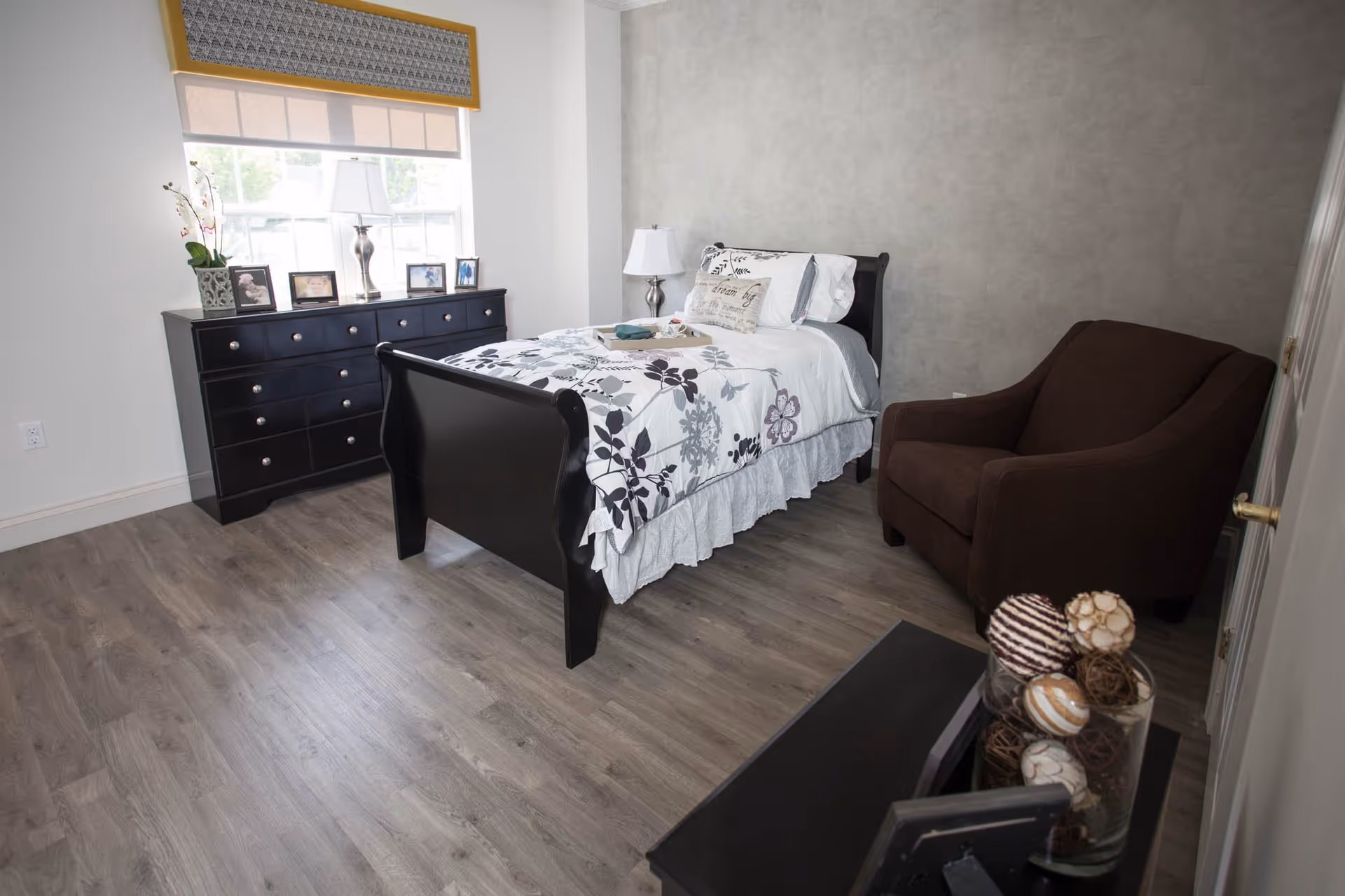A bedroom with a single bed featuring floral bedding, a dark wooden dresser with framed photos and a lamp on top, a brown upholstered armchair, and a window with a yellow valance letting in natural light. The floor is wood laminate and the walls are painted light gray and white.