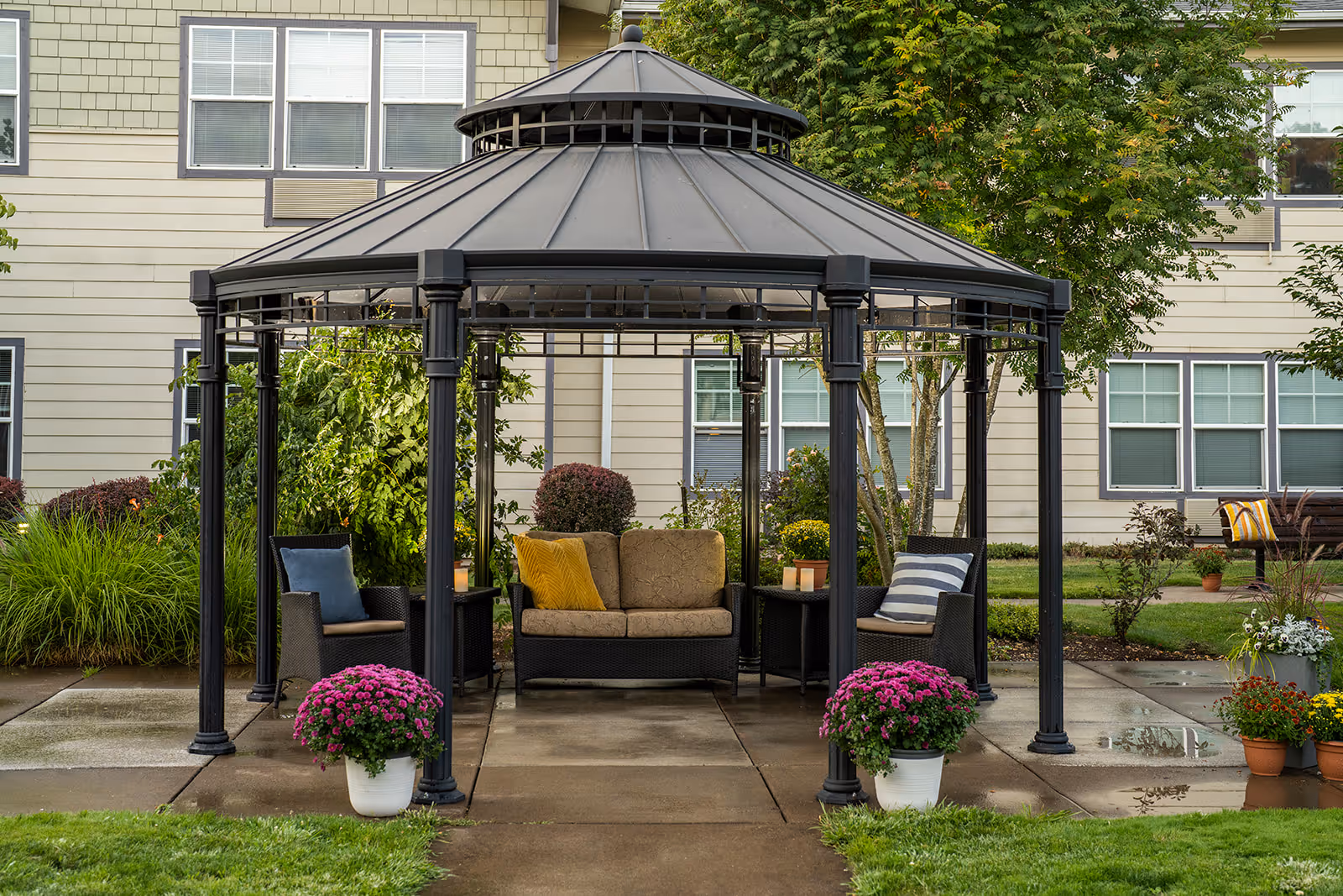 Outdoor gazebo with black metal frame and roof, furnished with cushioned wicker chairs and a loveseat with colorful pillows, surrounded by potted flowers and greenery, in front of a beige building with multiple windows.