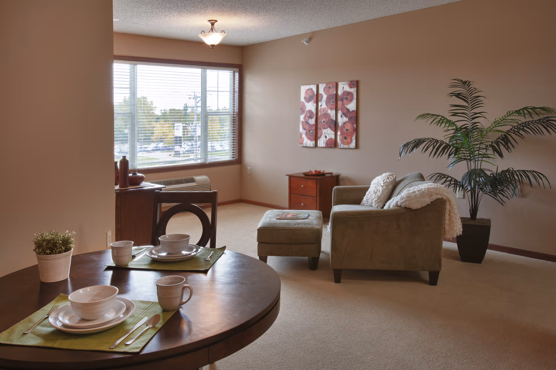 A cozy senior living room with a small round dining table set for two with white dishes and green placemats. In the background, there is a green armchair with a matching ottoman, a small wooden cabinet, a large potted plant, and a three-panel floral wall art. A large window with blinds lets in natural light.