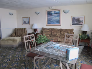 A cozy living room area with a patterned carpet, a glass table with a puzzle in progress, four chairs around the table, a floral-patterned sofa, an armchair with an ottoman, two side tables with lamps, and three framed landscape paintings on the wall.
