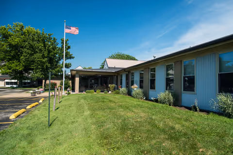 Front exterior of a single-story senior living facility with a flagpole, grassy lawn, and entrance under a blue sky.
